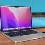 Open MacBook on wooden table with colorful abstract wallpaper on screen, blue cushion in background.