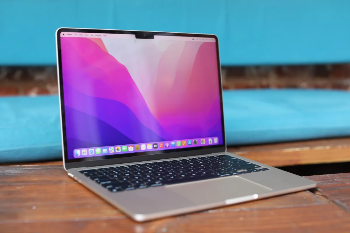 Open MacBook on wooden table with colorful abstract wallpaper on screen, blue cushion in background.