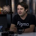 Person wearing a Figma shirt sits at a table with a Bloomberg mug, bookshelf and blurred background visible.