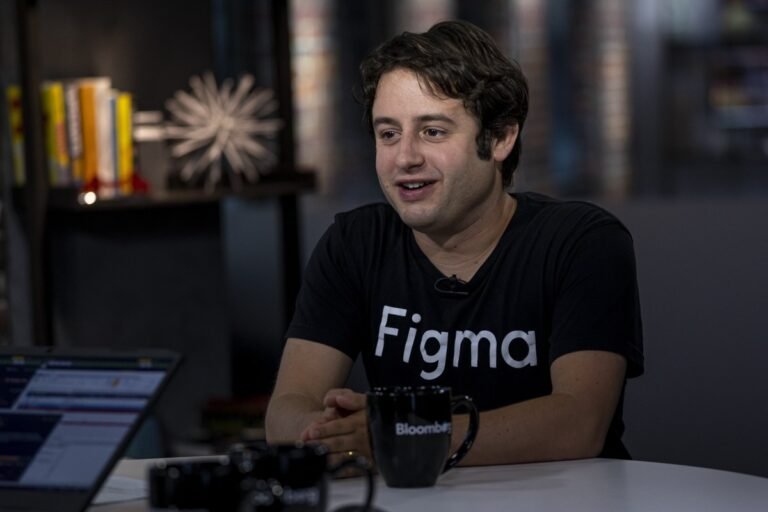 Person wearing a Figma shirt sits at a table with a Bloomberg mug, bookshelf and blurred background visible.
