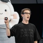 Man in black shirt holding a device speaks on stage, large cartoon figure in background, shirt text reads "building is my...