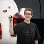 Man in glasses holding a small gadget, wearing a shirt with text "Building is my love language," large plush character in...
