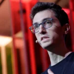 Person wearing glasses speaking on stage with a microphone, colorful geometric background.