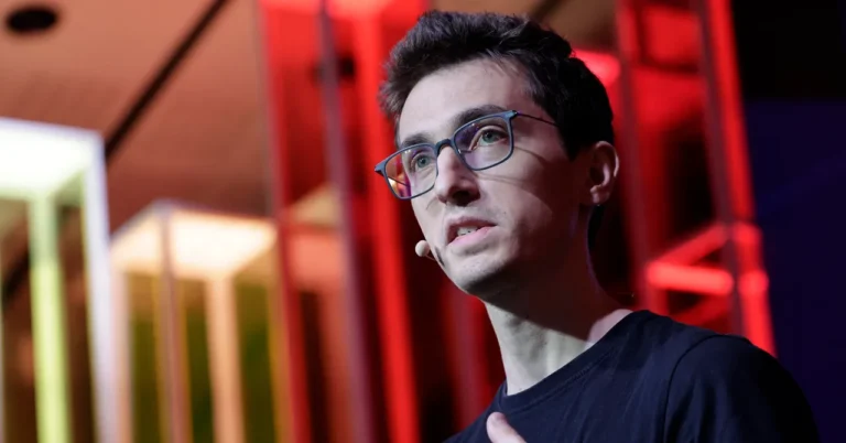 Person wearing glasses speaking on stage with a microphone, colorful geometric background.