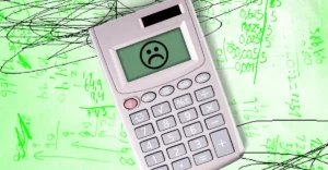 Calculator with a sad face on screen over green paper filled with handwritten mathematical equations and scribbles.
