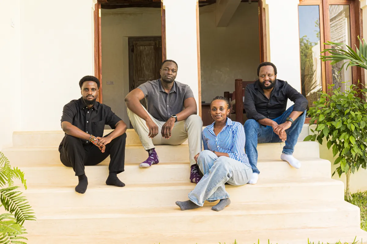 Four people sitting on outdoor steps in front of a building, surrounded by plants, looking at the camera.