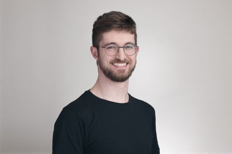 Man with glasses and beard smiling, wearing a black shirt, against a plain light background.