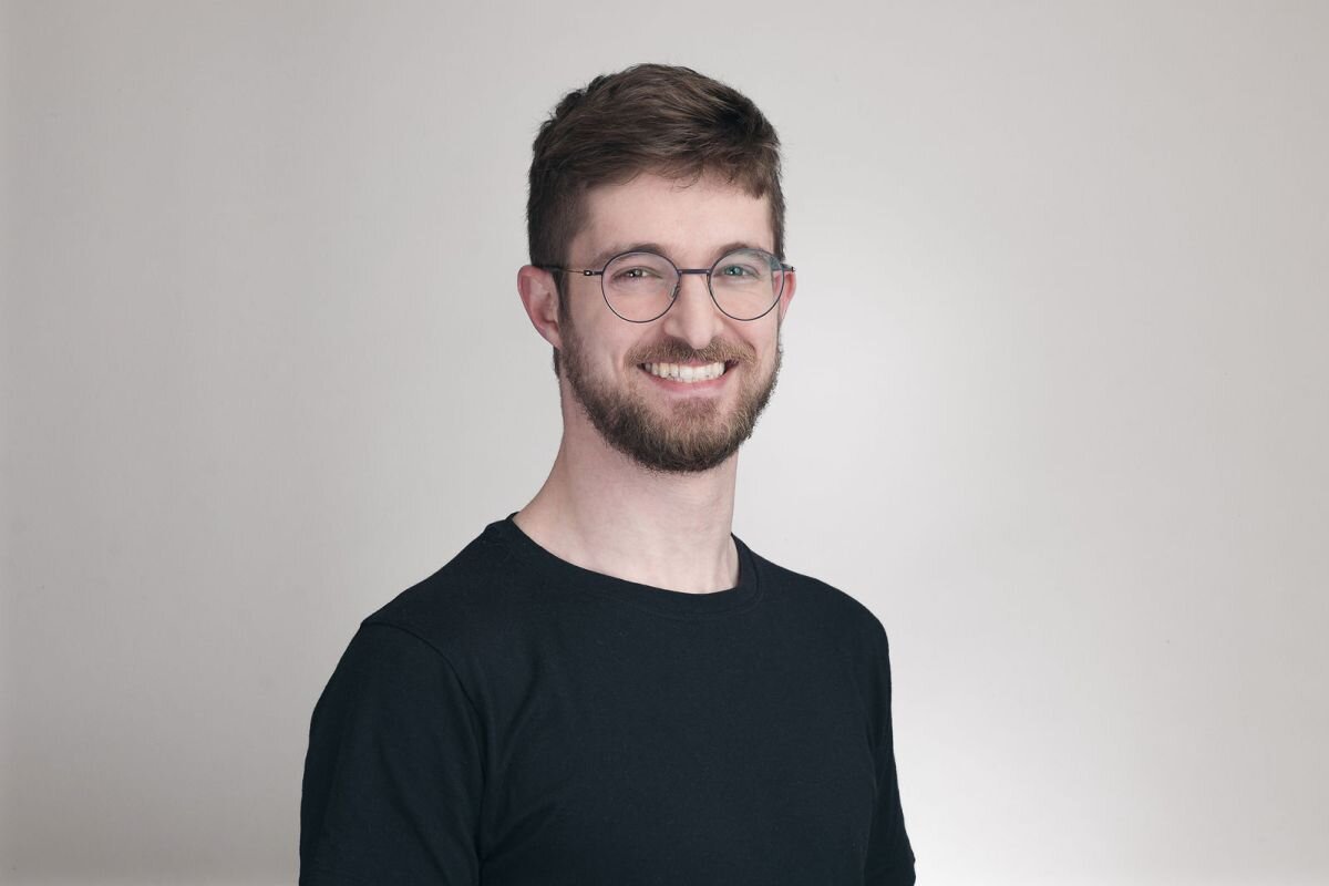 Man with glasses and beard smiling, wearing a black shirt, against a plain light background.