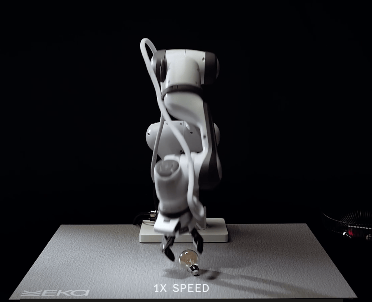 Robotic arm with a gripper holding a small object on a table, dark background, text "1X SPEED" and "EKA" visible