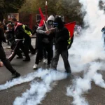 Protesters in hoodies and masks run through tear gas, some carrying red flags, on a city street with trees in the background.