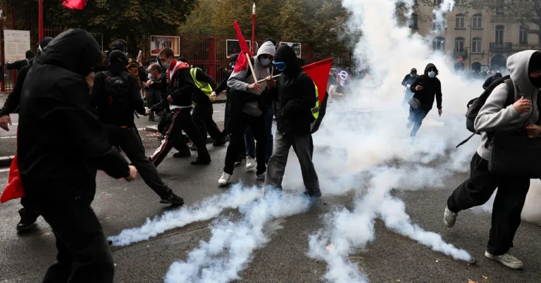 Protesters in hoodies and masks run through tear gas, some carrying red flags, on a city street with trees in the background.