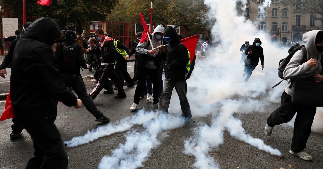 Protesters in hoodies and masks run through tear gas, some carrying red flags, on a city street with trees in the background.