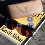 Sunglasses and a beige Ray-Ban case rest on a book titled "Kaveh Akbar" on a wooden surface.