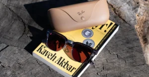 Sunglasses and a beige Ray-Ban case rest on a book titled "Kaveh Akbar" on a wooden surface.