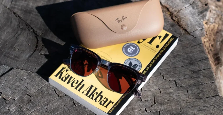 Sunglasses and a beige Ray-Ban case rest on a book titled "Kaveh Akbar" on a wooden surface.