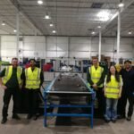 Five people in high-visibility vests stand in a warehouse next to industrial equipment, smiling at the camera.