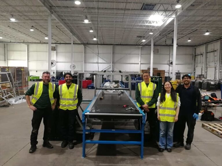 Five people in high-visibility vests stand in a warehouse next to industrial equipment, smiling at the camera.