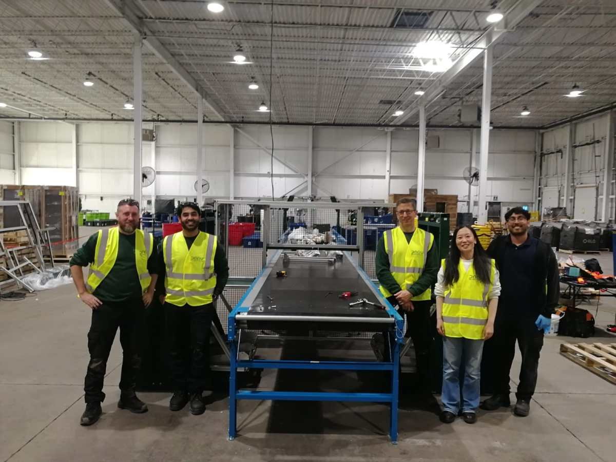 Five people in high-visibility vests stand in a warehouse next to industrial equipment, smiling at the camera.