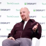 Bald man in maroon jacket speaks on stage at TechCrunch Disrupt with branded backdrop, water bottle on table.