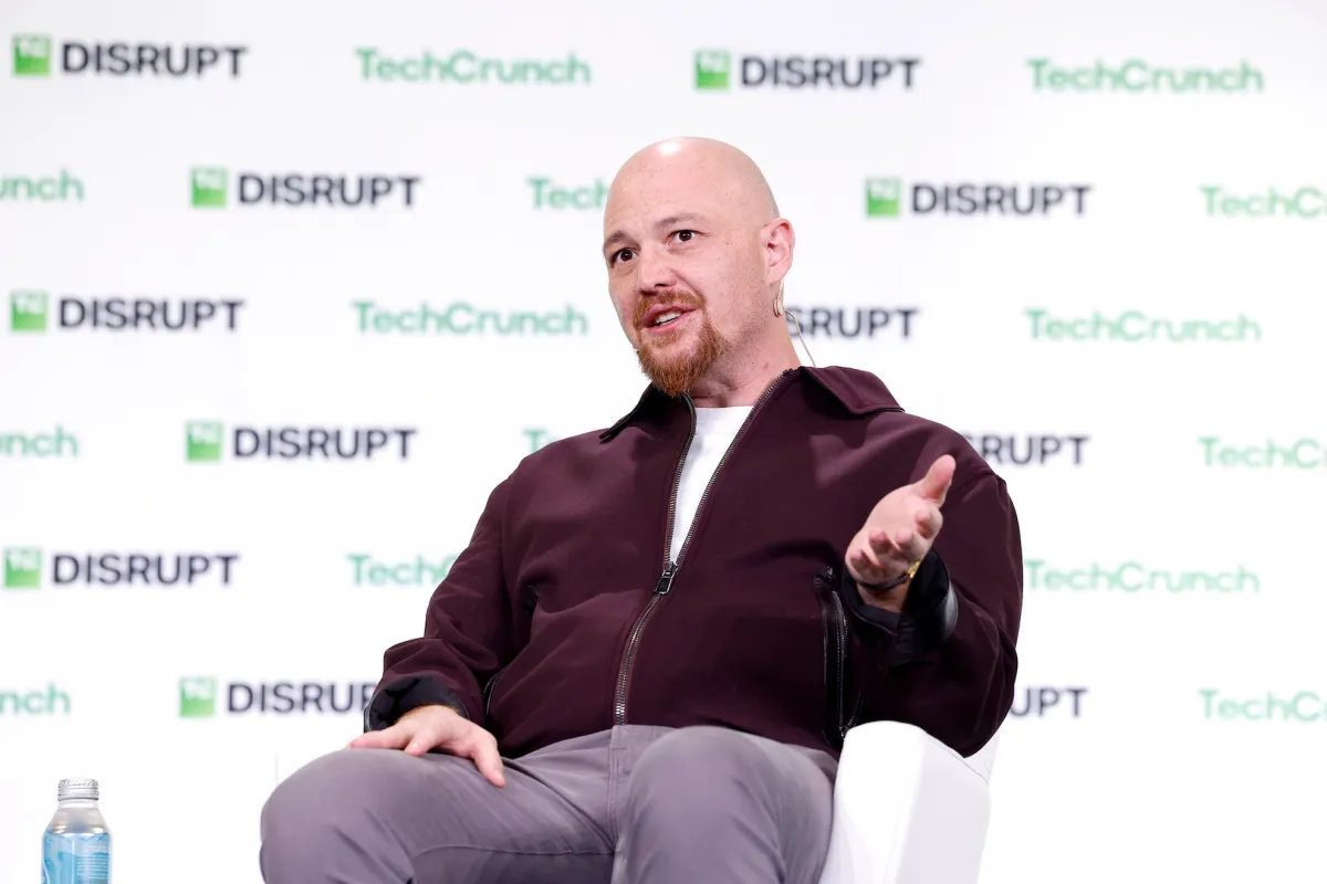 Bald man in maroon jacket speaks on stage at TechCrunch Disrupt with branded backdrop, water bottle on table.