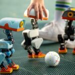 Three small, colorful robots on a grid floor near a white ball, with a hand adjusting one robot and legs wearing striped s...