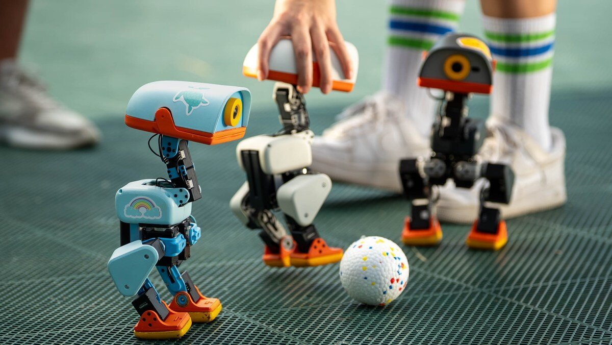 Three small, colorful robots on a grid floor near a white ball, with a hand adjusting one robot and legs wearing striped s...