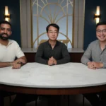 Three men sit at a round table in a modern room with geometric wall design and sconce lighting.