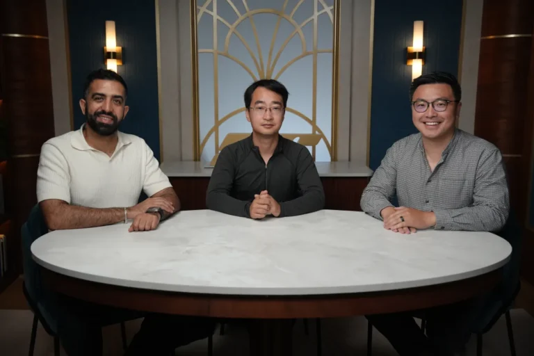Three men sit at a round table in a modern room with geometric wall design and sconce lighting.