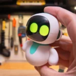 Hand holding a small, white robot with glowing green eyes, set against a blurred workshop background with tools.