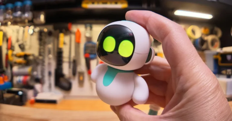 Hand holding a small, white robot with glowing green eyes, set against a blurred workshop background with tools.