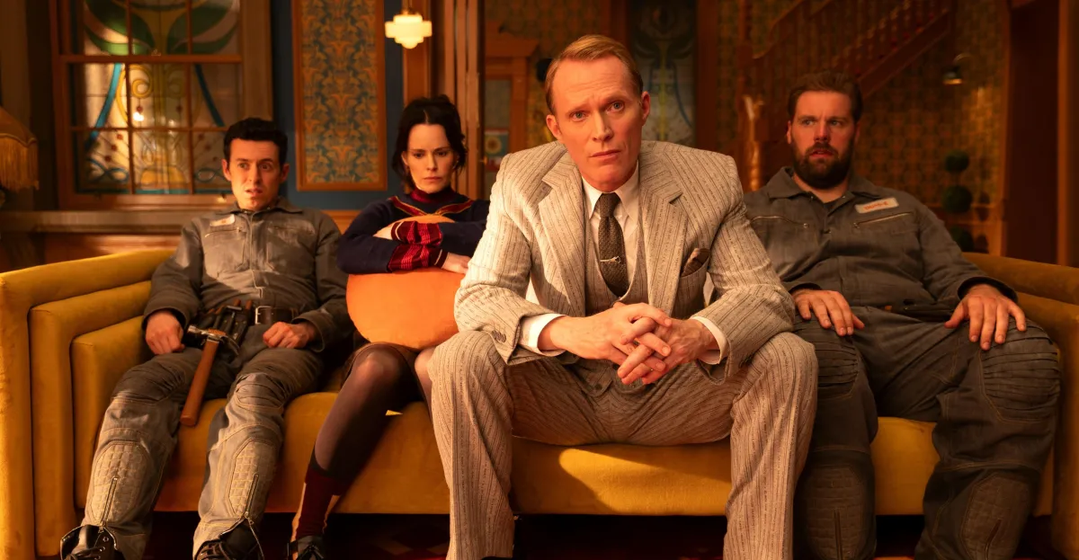 Four people sitting on a yellow couch in a vintage room; one in a suit, others in casual attire, looking intently ahead.