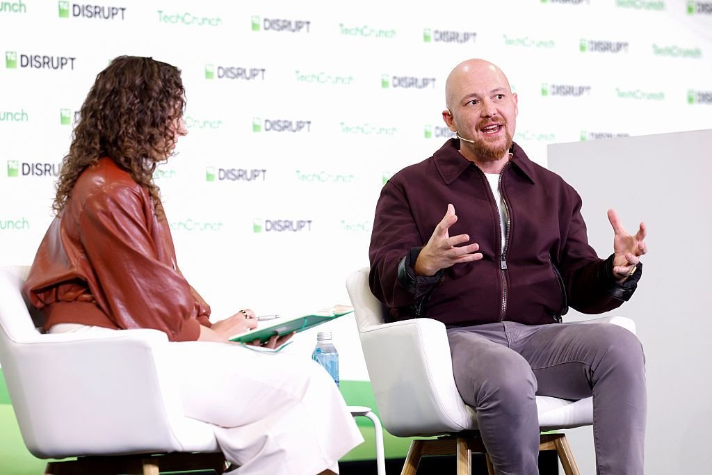 Two people seated on stage at TechCrunch Disrupt, engaged in conversation, with event branding in the background.