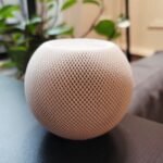 Small, spherical smart speaker with mesh exterior on a table, blurred background with plants and furniture.