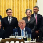 Group of executives and a central figure seated at a table in a formal setting with a yellow curtain background.