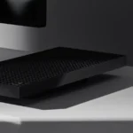 Sleek black rectangular device with textured surface on a gray desk, partially obscured monitor in the background.