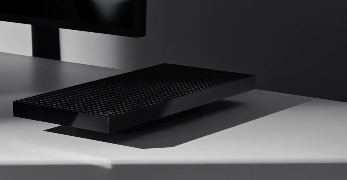 Sleek black rectangular device with textured surface on a gray desk, partially obscured monitor in the background.