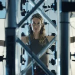 Woman with outstretched arms stands inside a structure surrounded by cameras and lights, looking directly ahead.