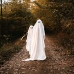 Two people in white ghost costumes walk down a leafy forest path, surrounded by green foliage.