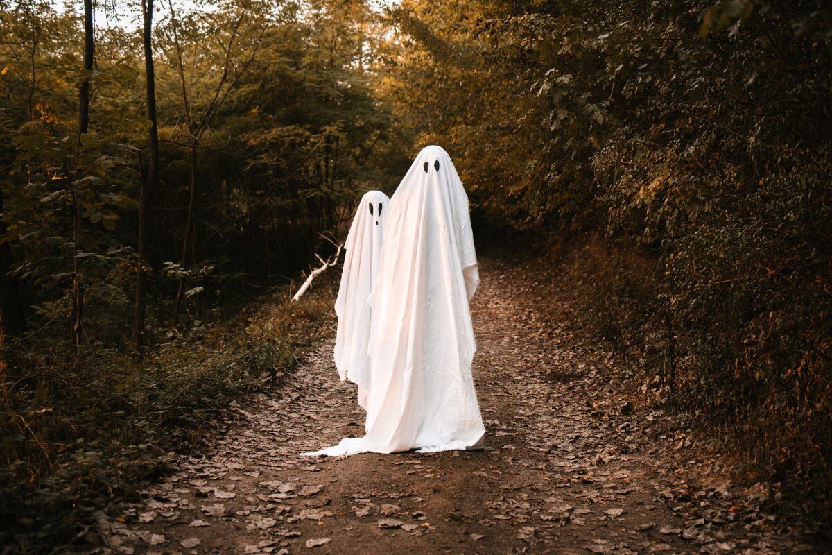 Two people in white ghost costumes walk down a leafy forest path, surrounded by green foliage.