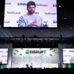 Speaker on stage at TechCrunch Disrupt with large screens displaying his image, audience watching in a dimly lit venue.