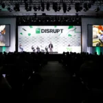 TechCrunch Disrupt stage with speakers, large screens displaying technology images, and audience in darkened auditorium.