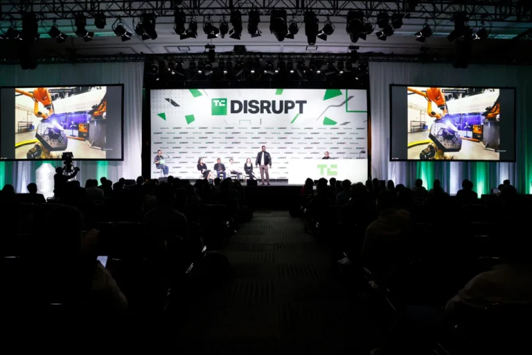 TechCrunch Disrupt stage with speakers, large screens displaying technology images, and audience in darkened auditorium.