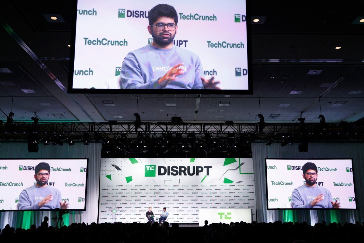 Speaker on stage at TechCrunch Disrupt with large screens displaying his image, audience watching in a dimly lit venue.