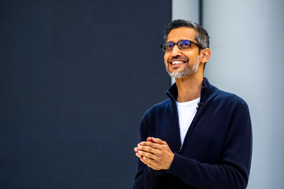 Smiling man in glasses and a dark sweater speaks on stage with a microphone, standing against a plain dark background.