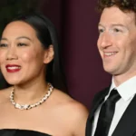 A man and woman smiling, both dressed in formal attire; woman in a black dress with a necklace, man in a black suit and tie.
