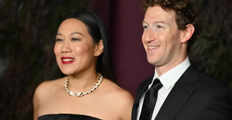 A man and woman smiling, both dressed in formal attire; woman in a black dress with a necklace, man in a black suit and tie.