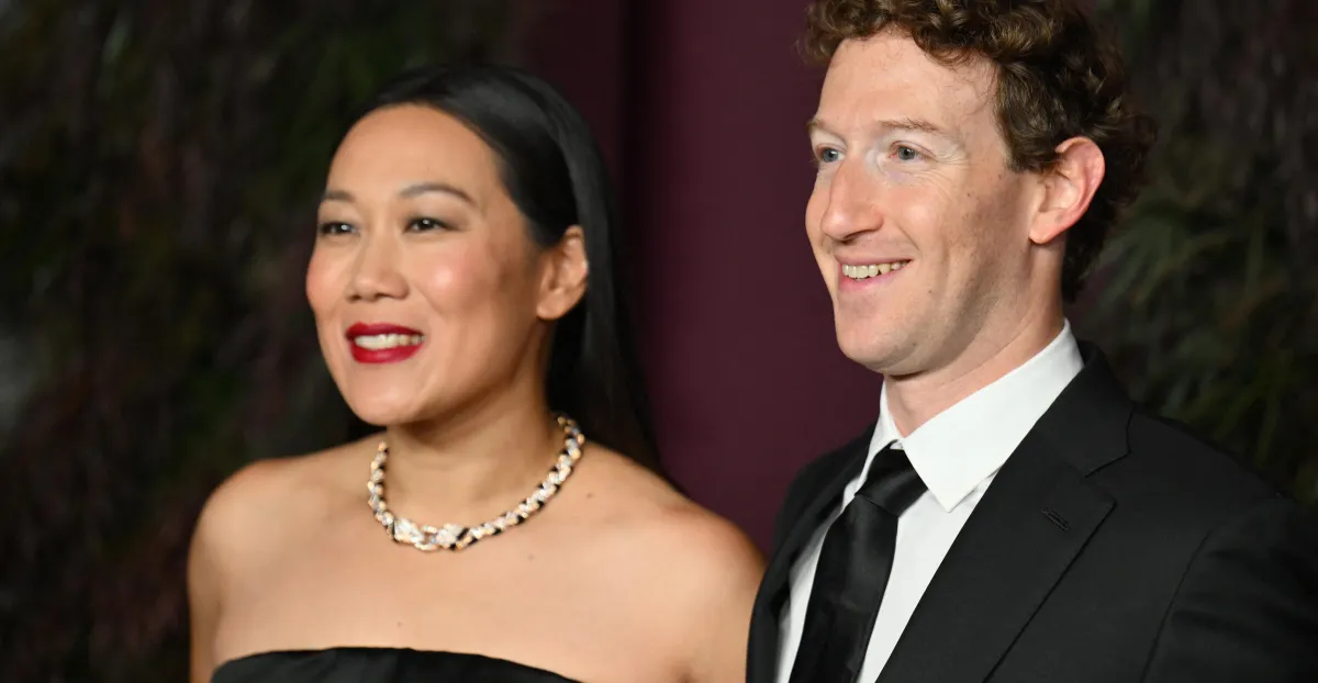 A man and woman smiling, both dressed in formal attire; woman in a black dress with a necklace, man in a black suit and tie.