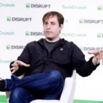 Person speaking on stage at TechCrunch Disrupt event, seated in a white chair, gesturing with hands, water bottle nearby.