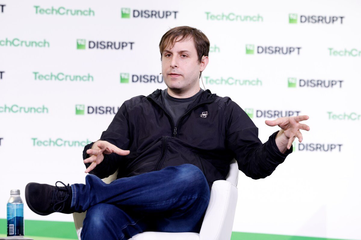 Person speaking on stage at TechCrunch Disrupt event, seated in a white chair, gesturing with hands, water bottle nearby.