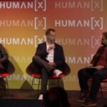 Three men seated on stage discussing at Human[X] event, with branded backdrop and one holding a notepad.